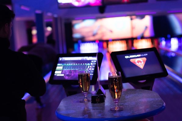 Två ölglas på ett litet bord, i bakgrund är bowlingbanor, dunkelt med blå och lila partybelysning.