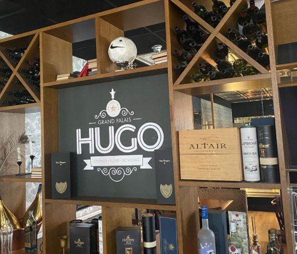 Brun trähylla med flaskor, på en hylla ligger en vit fotboll. Hugos logotyp i mitten.