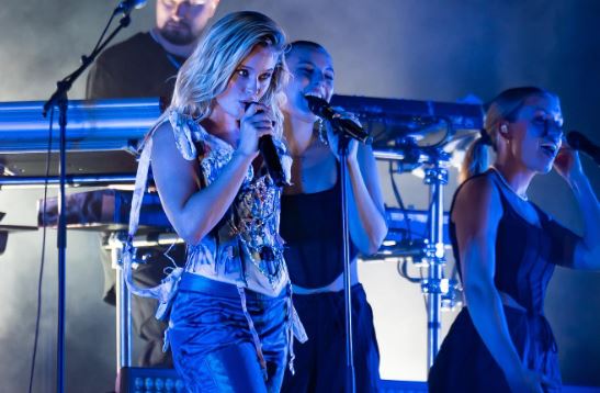 Zara i blått ljus med mikrofon och två dansare bakom.