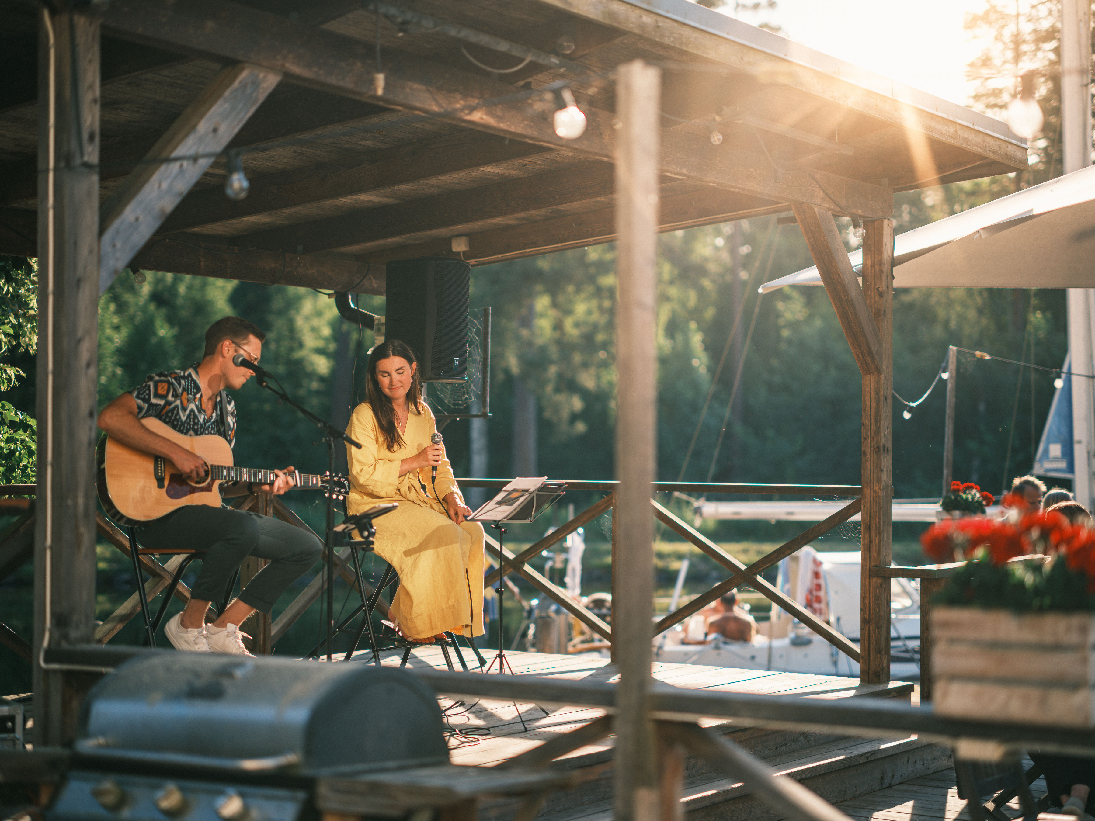 En solbränd person sitter på stol och spelar gitarr, bredvid person i långt mörkt hår och gul klänning som håller i en mikrofon. Båtar i bakgrunden. Solsken.